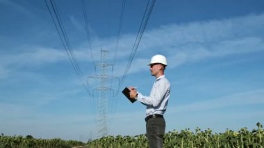 Beyaz kasklı bir adam, yüksek voltajlı elektrik hattının yanındaki tarlada dururken tablet üzerinde çalışır. Güç şebekesi durumu izleme.