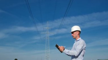 Elektrik mühendisi bir tablet üzerinde çalışır, yüksek voltajlı bir elektrik hattının ve mavi bir gökyüzünün arka planında durur, kameraya doğru döner ve tableti aşağı indirir..