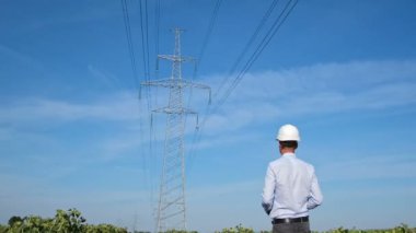 Beyaz kasklı bir adam yüksek voltajlı elektrik hattı boyunca yürür ve metal bir direğe ulaşır. Kamera arkadan bir adam yakalar..