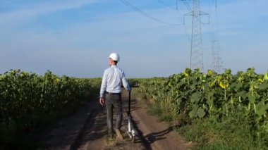 Beyaz kasklı bir adam, ayçiçeği tarlaları arasında asfaltsız bir yolda yürüyor ve yanında elektrikli bir scooter yuvarlıyor..