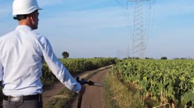 Beyaz korumalı miğferli bir adam, yüksek voltajlı elektrik hattı boyunca ayçiçeği tarlaları arasında yürürken yanına elektrikli scooter takar ve kamera adamı arkadan yakalar..