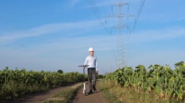 Beyaz korumalı miğferli bir mühendis, ayçiçeği tarlaları arasında yol boyunca yürür ve yakınlarda bir elektrikli scooter yuvarlar, bir adam video kameraya doğru yürür..