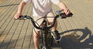 Çocuk yavaşça bisiklet zili çalarak gidona odaklanıyor. Güneşli caddede sürerken çocuk direksiyonu dikkatle kontrol ediyor.