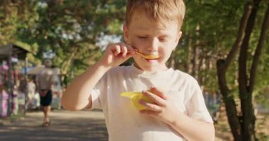 Sarışın çocuk sarı tatlı kabını tutuyor ve küçük kaşıkla dondurma yiyor. Çocuk porsiyonunu kaldırır dikkatlice çiğner. Lezzetli yiyecekler tüm dikkatle park alanında dinlenir.