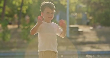 Anaokulu çocuğu parlak güneş ışığına bakarken kollarını enerji ile sallıyor. Çocuk tekrar tekrar elini kaldırıyor ve şehir parkının çevresini neşeli hareketlerle selamlıyor.