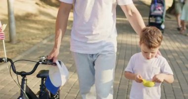 Babam küçük bir çocukla yürürken sağ eliyle park yolu boyunca bisikleti itiyor. Oğlu dondurma yerken yavaş yavaş lezzetli tatlılara odaklanıyor.