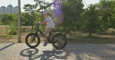 Anaokulu çocuğu kaldırım parkında kendine güvenerek bisiklete biniyor. Genç çocuk hızla pedal çevirirken ağaçlara bakıyor.