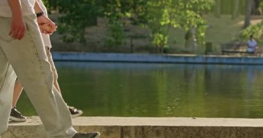 Sorumluluk sahibi baba, göletin kenarında dururken küçük çocuğu eliyle tutuyor. Erkekler, su bariyerinde yürürken dengeyi korumak için anaokulu çocuklarını destekliyor.