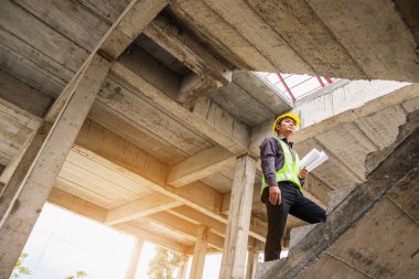 Genç profesyonel mühendis koruyucu miğfer ve bina şantiyesinde elinde proje kağıtları ile çalışıyor.