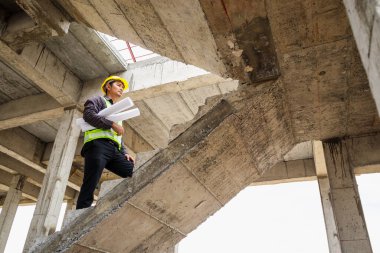 Genç profesyonel mühendis koruyucu miğfer ve bina şantiyesinde elinde proje kağıtları ile çalışıyor.