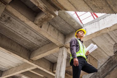 Genç profesyonel mühendis koruyucu miğfer ve bina şantiyesinde elinde proje kağıtları ile çalışıyor.