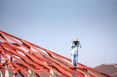 İnşaat işçileri binanın çatısının çelik iskelet yapısını inşa ediyorlar. İnşaat alanında bulutlar ve gökyüzü var.