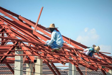 İnşaat işçileri binanın çatısının çelik iskelet yapısını inşa ediyorlar. İnşaat alanında bulutlar ve gökyüzü var.