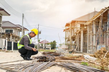 Asyalı işadamı inşaat mühendisi koruyucu miğfer ve bina inşaatındaki çelik çubuklara bakan bina planı kağıtları.