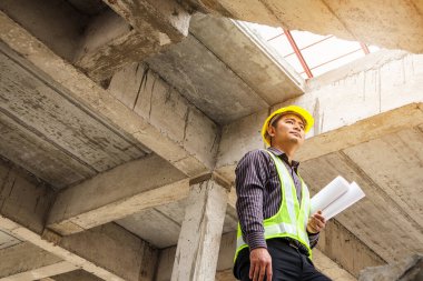 Genç profesyonel mühendis koruyucu miğfer ve bina şantiyesinde elinde proje kağıtları ile çalışıyor.