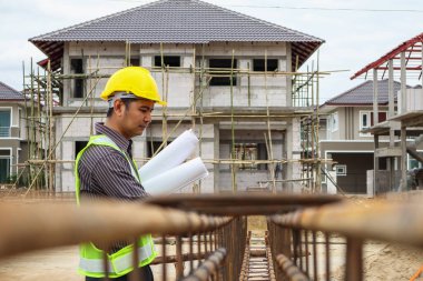 Asyalı işadamı inşaat mühendisi koruyucu miğfer ve bina inşaatındaki çelik çubuklara bakan bina planı kağıtları.