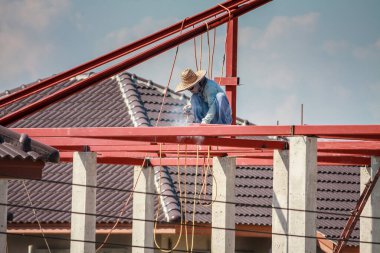 İnşaat işçileri binanın çatısının çelik iskelet yapısını inşa ediyorlar. İnşaat alanında bulutlar ve gökyüzü var.