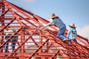 İnşaat işçileri binanın çatısının çelik iskelet yapısını inşa ediyorlar. İnşaat alanında bulutlar ve gökyüzü var.