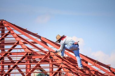 İnşaat işçileri binanın çatısının çelik iskelet yapısını inşa ediyorlar. İnşaat alanında bulutlar ve gökyüzü var.