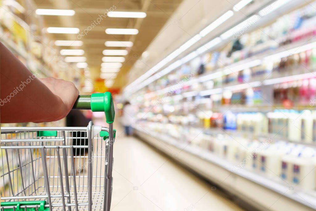 woman hand hold supermarket shopping cart with abstract blur refrigerator shelves with fresh milk bottles and dairy products in grocery store