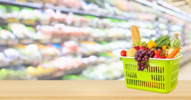 Tahta masanın üzerinde meyve ve sebzelerle dolu alışveriş sepeti. Marketin arka planında bulanıklık var.