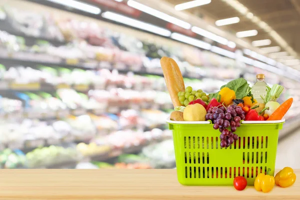 Tahta masanın üzerinde meyve ve sebzelerle dolu alışveriş sepeti. Marketin arka planında bulanıklık var.