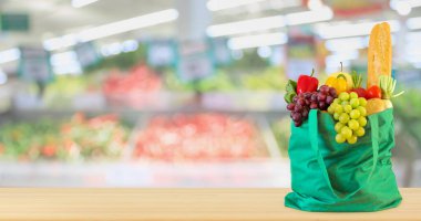 Yeniden kullanılabilir yeşil alışveriş poşetinde taze meyve ve sebzeler ahşap masanın üstünde süpermarket bakkalının bokeh ışıklı bulanık arka planıyla