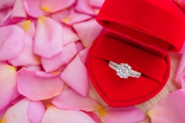 Kırmızı kalp mücevher kutusunda zarif bir alyans. Güzel pembe gül yapraklı arka planda. Yakın plan.