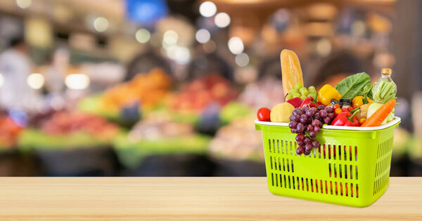 Green shopping basket with groceries product on wood table top with abstract blur colorful Fruits in display basket in supermarket grocery store defocused background with bokeh light