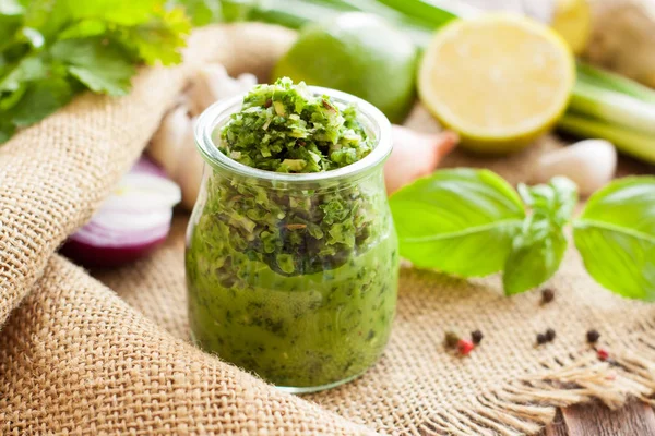 Green sauce in a glass jar in background with basil, lime, onion and parsley. Thai green curry paste