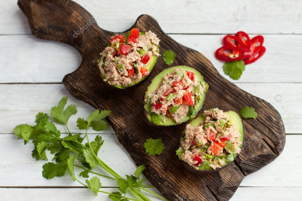 Avocado appetizers stuffed with canned tuna, bell pepper, herbs on wooden cutting board