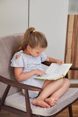 Küçük sarışın kız odasında rahat yumuşak sandalyede otururken hikaye kitabı okuma