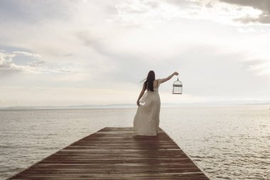 Uzun beyaz elbiseli zarif bir kadın elinde boş bir kafesle denize doğru yürüyor..