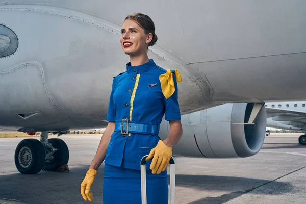 Радостная стюардесса с багажом, стоящим у самолета — стоковое фото