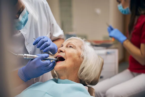 Мужчина-дантист с зубными инструментами и дрелью во время ухода за зубами пациента в офисе стоматологической клиники — стоковое фото