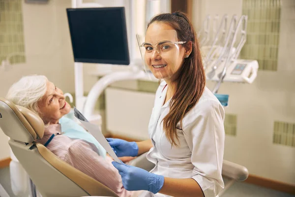 Χαμογελαστή γυναίκα οδοντίατρος με λευκή στολή κρατώντας φωτογραφία του πελάτη δόντια — Φωτογραφία Αρχείου