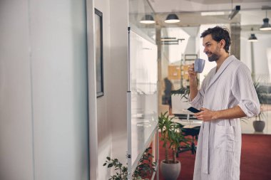 Bornozlu neşeli genç adam iş yerinde kahve içiyor.