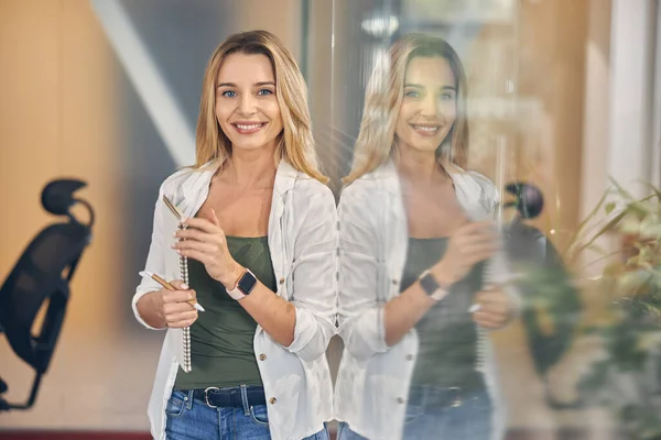 Радісна молода жінка стоїть біля скляної стіни в офісі — стокове фото