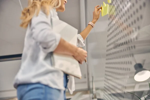 Kvinnlig kontorsarbetare sätta klibbig anteckning på glasvägg — Stockfoto