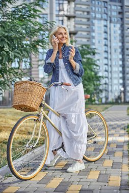 Uzun beyaz elbiseli ve kot ceketli güzel bir bayan açık havada eski bir bisikletle yürüyor.