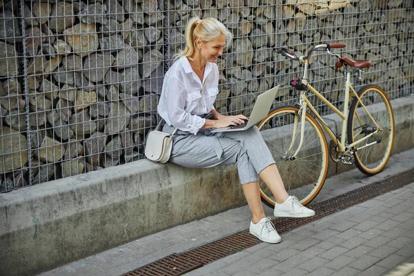 Fashionabel glad kvinna i affärer casual kläder sitter framför stenvägg med bärbar dator — Stockfoto