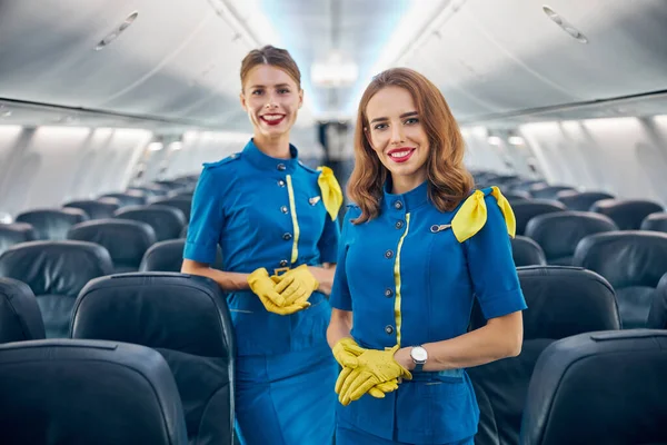 Dos mujeres auxiliares de vuelo sonriendo a la cámara fotográfica en el salón de aviones de pasajeros —  Fotos de Stock