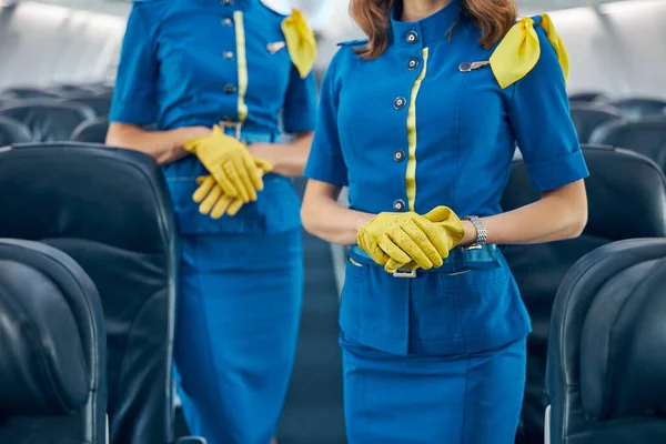 Beautiful two air hostesses in yellow gloves standing in aisle of board passenger aircraft — Stock Photo, Image