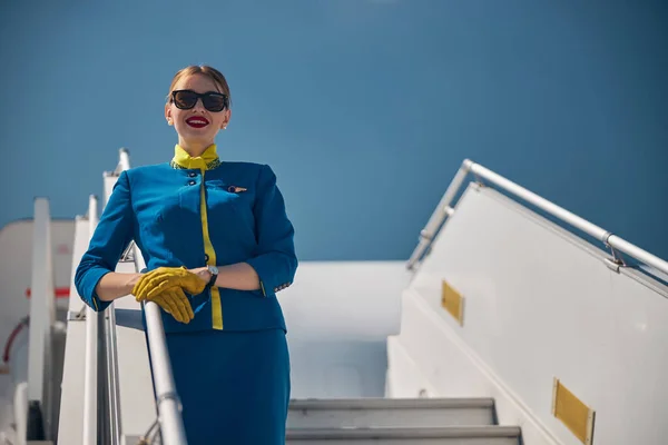 Веселая стюардесса, стоящая на ступенях самолета под голубым небом — стоковое фото