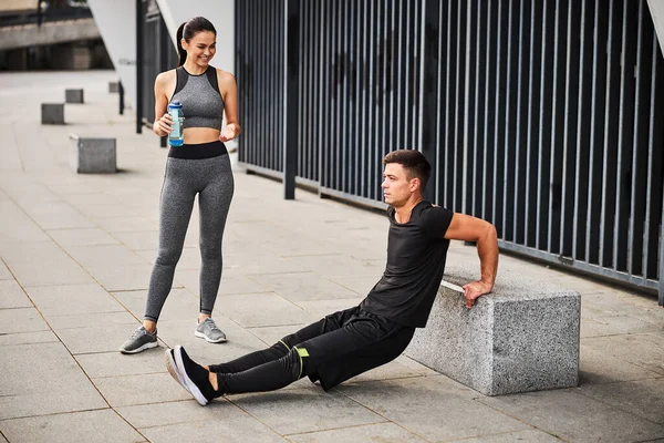 Jolly young woman assisting boyfriend during training outdoors — Stock Photo, Image