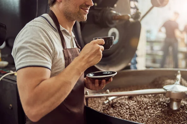 Sonriente trabajador masculino bebiendo café cerca de máquina de tostado de café —  Fotos de Stock