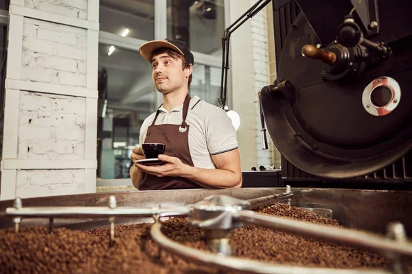 Hombre guapo con bebida caliente de pie por máquina de tostado de café —  Fotos de Stock