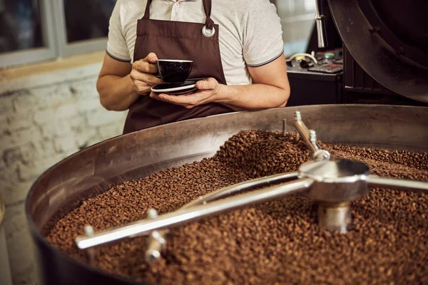 Trabajador masculino con bebida caliente de pie por máquina de tostado de café —  Fotos de Stock