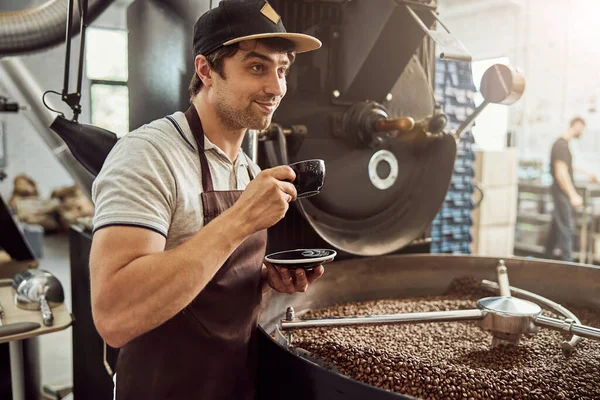 Cheerful man roaster drinking coffee near coffee roasting machine — Stock Photo, Image