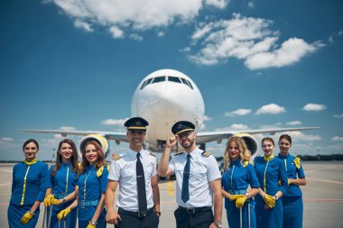 Neşeli havayolu işçileri mavi gökyüzünün altında havaalanında duruyorlar.